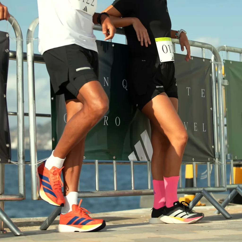 Foto em plano médio mostrando as pernas de um homem e uma mulher, ambos corredores, encostados em uma grade de metal com banners de um evento de corrida. O homem, à esquerda, usa tênis Adidas Adizero laranja e azul. A mulher, à direita, usa tênis Adidas Adizero preto e branco com listras coloridas e meias rosa vibrantes, com um número de peito "017" visível no shorts. O fundo mostra a água do mar sob um céu azul ensolarado.