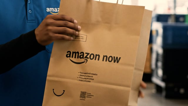 Fotografia de um entregador vestindo o uniforme azul da empresa segurando uma sacola de papel kraft com o logotipo impresso, ilustrando como as compras do Amazon Now no Brasil são preparadas para a entrega rápida.
