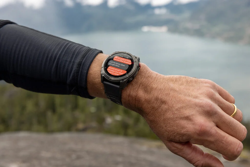Braço de uma pessoa ao ar livre usando o smartwatch Garmin Fenix 8 Pro. A tela do relógio exibe notificações de mensagens de texto, com uma paisagem de montanhas e um lago desfocados ao fundo.