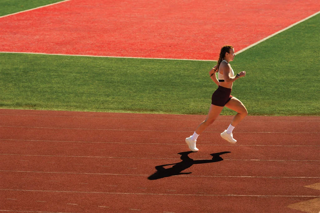 Atleta feminina correndo em uma pista de atletismo vermelha em um dia ensolarado, utilizando roupas esportivas e um smartwatch Garmin Forerunner 970 no pulso para registrar o treino um dos melhores smartwatches Garmin