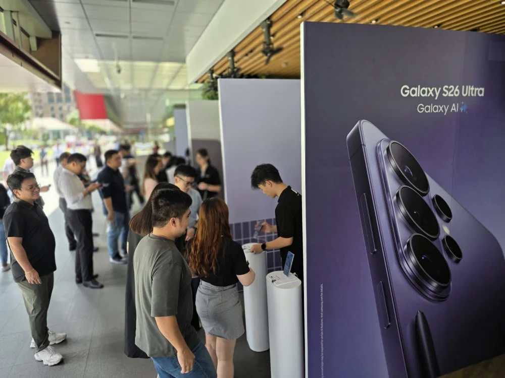 Fotografia de um estande promocional da Samsung com pessoas interagindo em totens. Ao lado direito, há um grande painel roxo exibindo a traseira e as câmeras do Galaxy S26 Ultra com o logotipo "Galaxy AI". Este dispositivo será um dos principais beneficiados pelas novas integrações de inteligência artificial da One UI 9 e Android 17.
