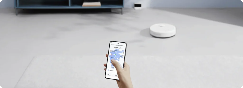 Uma mão segurando um smartphone que exibe um mapa azul no aplicativo, enquanto um robô aspirador xiaomi branco aparece ao fundo em um piso claro.