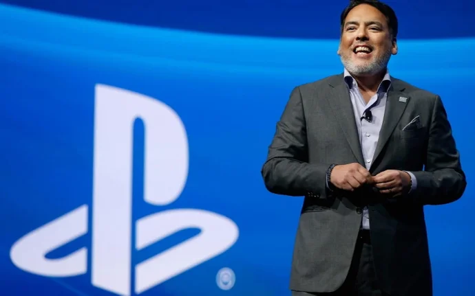 Fotografia de Shawn Layden, ex-CEO da PlayStation, sorrindo e discursando em um palco com um grande logo da PlayStation ao fundo em um telão azul.